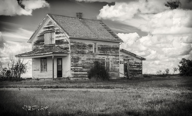 horizontal black and white image of an old rustic abandoned house with a veranda sitting under a cloudy sky.