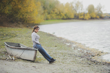The girl sitting on the edge of the old boat near the shore of the river. Look into the camera. Autumn.