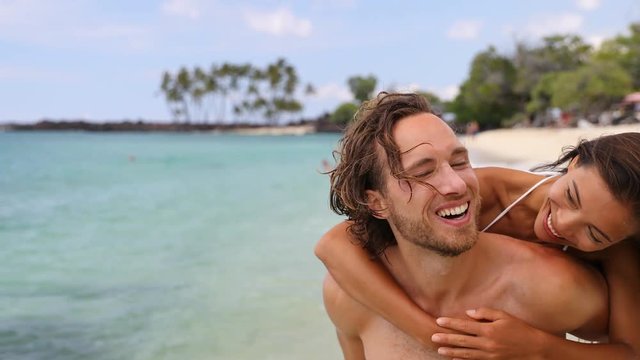 Attractive happy couple laughing having fun piggybacking on tropical beach. Caucasian man carrying Asian girlfriend having fun laughing on travel vacation. Healthy happy interracial people. 59.94 FPS.