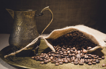 Vintage still life with heap of coffee beans in burlap sack near old copper turkish coffee pot on bronze tray against canvas background