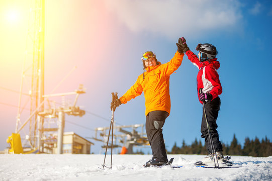 Man and woman holding hands, smiling looking at each other standing with skis on mountain top and giving each other a high five at a winter resort with ski lifts and blue sky in background