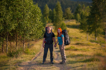Obraz premium Young tourists with backpacks holding hands on a trail to the mountain against trees and forest in the distance on the background