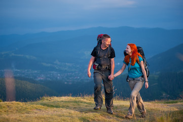 Obraz premium Young family of hikers with backpacks walking in the beautiful mountains area, holding hands and looking to each other. Lifestyle active vacations concept mountains landscape on background