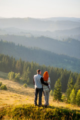 Fototapeta premium Charming couple tourists standing on a hill, enjoying beautiful mountain landscape with morning haze over the mountains and forests. Man hugging red-haired girl, she is covered with a plaid. Back view