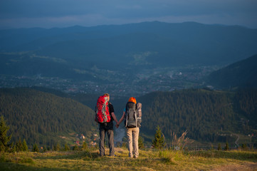 Rear view pair with backpacks holding hands standing on the ridge of hill, enjoying the view of beautiful open overlook on the mountains and village in the valley