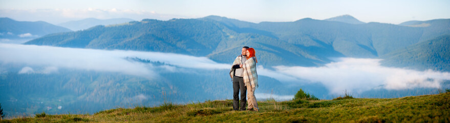 Fototapeta na wymiar Sweety couple hikers covered with a blanket standing together on a hill, enjoying beautiful mountain landscape with morning haze over the mountains and forests. Panorama
