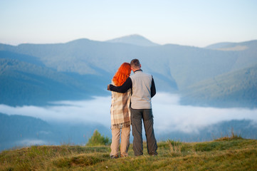 Romantic couple - girl and guy standing on a hill enjoying a morning haze over the mountains. Man hugging red-haired girl. Woman is covered with a plaid. Back view