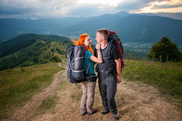 Loving couple hikers is embracing, standing with backpacks on the road in the mountains. Forests, hills and clouds sky on background