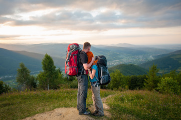 Romantic couple hikers with backpacks standing embracing and enjoying the view of beautiful open overlook on the mountains, forests, hills, village in the valley and cloudy sky