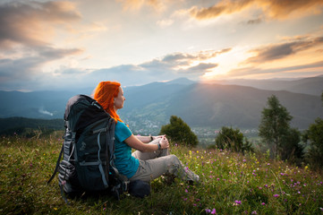 Smiling woman with backpack sitting in the grass with wild flowers on a hill and looking over the mountains, village in the valley and the rays of the sun breaking through the clouds at sunset