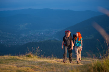 Tourists man and woman with backpacks walking in the beautiful mountains area, holding hands and looking to each other. Lifestyle active vacations concept mountains landscape on background