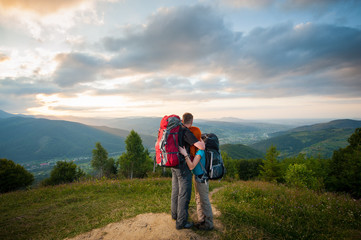 Fototapeta na wymiar Back view of couple tourists with backpacks standing embracing and enjoying the view of beautiful open overlook on the mountains, forests, hills, village in the valley and cloudy sky