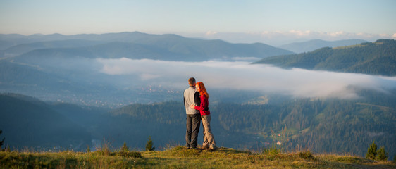 Romantic couple hikers hugging and enjoying beautiful mountain landscape with morning haze over the mountains and forests. Panorama