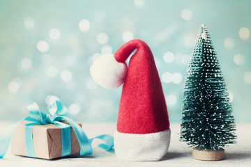 Miniature fir tree, santa hat and christmas gift box against blue bokeh background. Holiday...