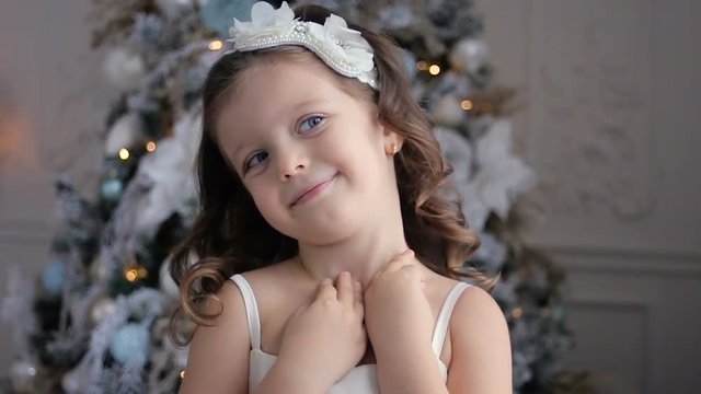 little girl three years old in a white dress smiling at the decorated Christmas tree in room