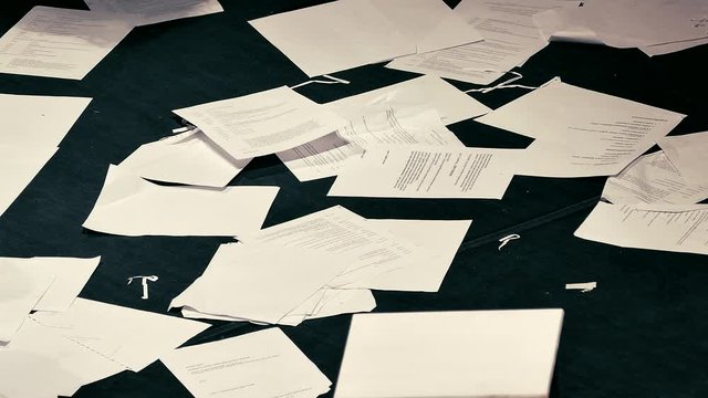 Sheets of paper with unreadable printed text fall to the black floor in a mess. People walk on the debris.
