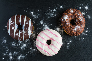 Chocolate and glazed donuts, pink, white, brown on black stone, top view.