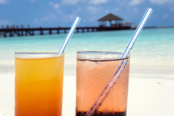 Two colorful cocktails laying on white sand, small corals covered beach in front of the amazing turquoise water of the indian ocean.