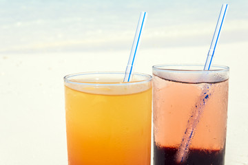 Two colorful cocktails laying on white sand, small corals covered beach in front of the amazing turquoise water of the indian ocean.