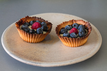 Two tartletes with custard, blueberries and raspberries on gray kitchen table. 