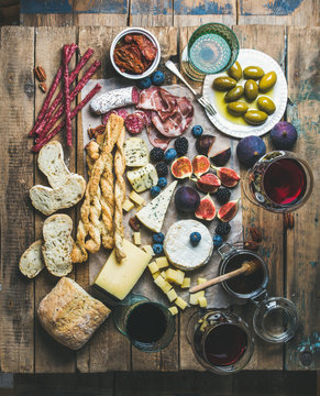 Wine and snack set with various wines in glasses, meat variety, bread, sun-dried tomatoes, honey, green olives, figs, nuts and fresh berries on wax paper over rustic wooden table background, top view