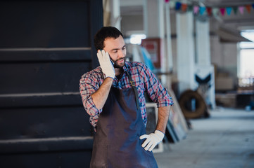 Fototapeta premium Smiling bearded man wearing gloves, owner of a small woodwork business, standing in his workshop studio, talking on mobile phone