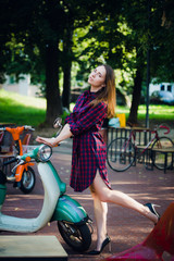 Obraz premium Urban fashion. Outdoor portrait of pretty young woman wearing checkered dress standing near scooter at park outdoors