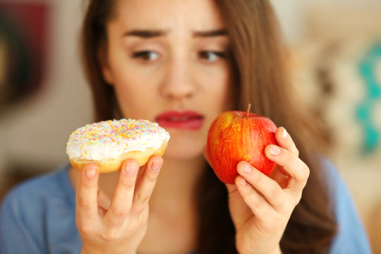 Beautiful young woman making choice between apple and donut, close up