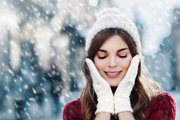 Outdoor close up photo of  young happy smiling girl walking on street. Model closed her eyes and touching face, wearing stylish winter hat and gloves. Christmas, New Year concept. Copy space for text