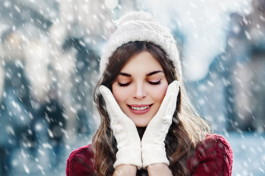 utdoor close up portrait of  young happy smiling girl walking on street. Model closed her eyes and touching face, wearing stylish winter hat and gloves. Magic snowfall. Christmas, New Year concept