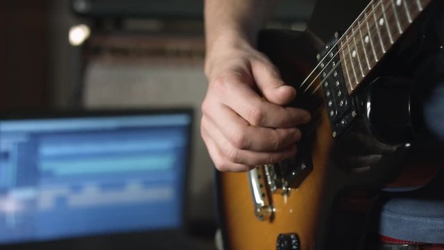 Close up of session musician playing electric guitar fingerpicking style and recording track with multitrack music software on laptop