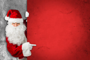 Santa claus hiding and looking through behind empty red concrete billboard  wall and pointing with...