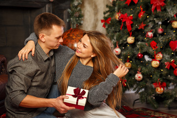 Fototapeta na wymiar Loving couple and Christmas. Man gives a girl a Christmas gift. They sit on the floor near the fireplace. In the background, a beautiful Christmas tree.