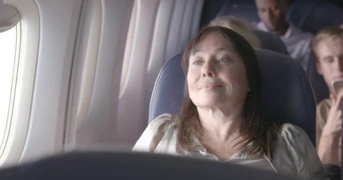 View across seats of mature woman sitting next to window and reclining her seat back to rest in main cabin of commercial airliner. Close up from front, recorded hand held at 60fps.