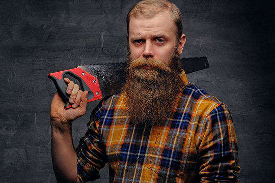 Bearded carpenter dressed in a plaid shirt holds handsaw.