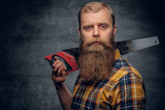 A bearded carpenter man dressed in a plaid shirt holds handsaw.
