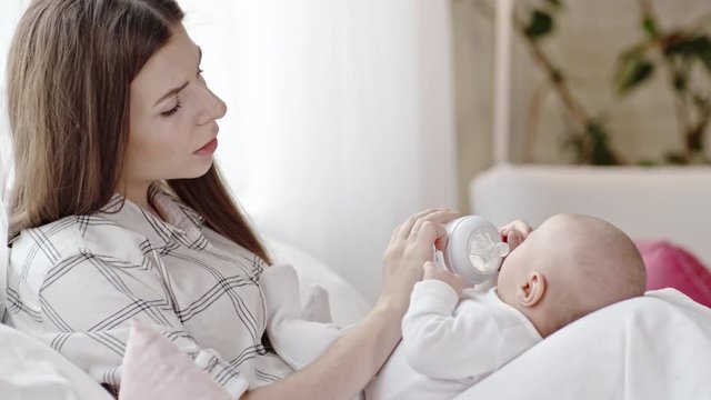 Young woman sitting with infant baby boy in her lap and giving him water in bottle