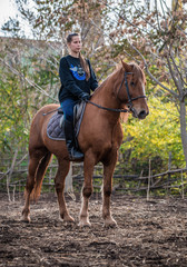 Young beautiful brunette girl rides a horse on a warm and sunny autumn day. Portrait of a pretty young woman on the horse, wearing tall boots and gloves.
