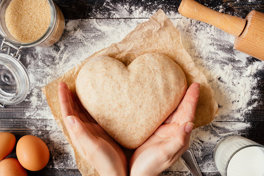 female hands holding dough in heart shape top view. Baking ingredients on the dark wooden table - Powered by Adobe