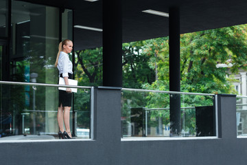 Fototapeta premium Young businesswoman holding laptop standing at the glass buildin