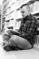 Handsome young student sitting on library floor reading book in