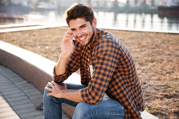 Cheerful young man sitting and talking on cell phone outdoors