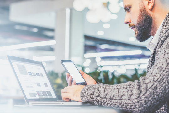 Side
 view of young bearded businessman,dressed in gray cardigan,sitting at 
table in cafe with modern interior and is holding smartphone while using
 laptop.Man uses gadget,blogging.Film effect.