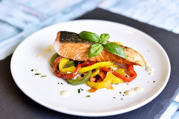 Delicious salmon filet and red bell pepper vegetables on a white plate. Healthy meal, decorated with basil leaf.