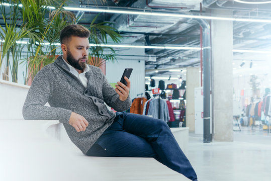 Side view.Young bearded businessman,dressed in white shirt and gray cardigan,sitting in room with a modern interior and uses the smartphone. In the background houseplants. A man uses the gadget.