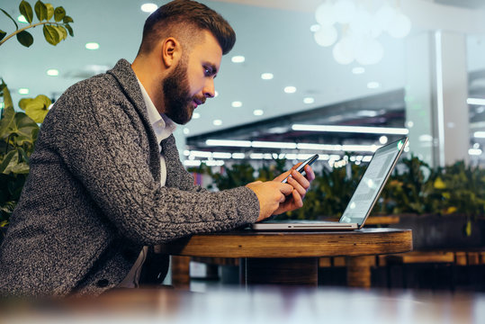 Side view of young bearded businessman,dressed in gray cardigan,sitting at round wooden table in cafe with modern interior and is holding smartphone.Laptop on table.Man uses gadget. Blurred background