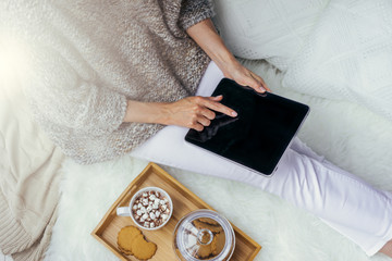 Top view of the girl touching screen tablet computer in her hands. The girl, dressed in a knitted sweater sitting on a white carpet. Next cup of cocoa with marshmallows and cookies in a glass jar.