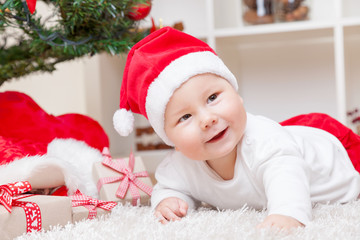 Cute baby in a Santa hat next to Christmas tree with presents