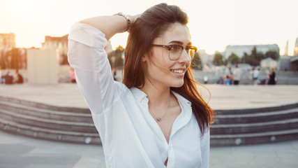 A cropped photo of a beautiful brunette woman in the glasses wearing white shirt. A charming girl with long brown hair is looking at the camera while standing on a city park background.