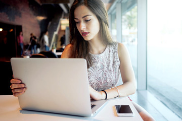 Young attractive woman is sitting in front of an open laptop in a coffee shop. Modern young business woman is working on a new project, looking at the screen of a portable computer.